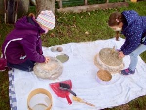 Ein großes Tischtuch ist auf einer Wiese ausgebreitet, darauf zwei Handmühlen: Ein Reibestein und eine Drehmühle. Zwei Kinder mahlen darauf Korn.