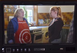 Ein Frau mit roter Schürze und eine Teenagerin stehen sich in einer Küche gegenüber, umrahmt von einem externen Kameramonitor