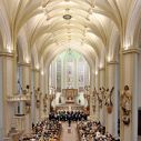 Blick von oben auf ein helles gotisches Kirchenschiff, die Bänke sind voll besetzt, im Altarraum steht ein Chor. Das weiße Gewölbe ist hell erleuchtet.
