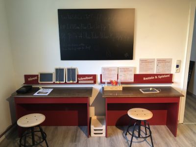 An einer Wand hängt eine schwarze Tafel, auf der das Alphabet in Sütterlin steht. Davor zwei rot gestrichene Tische mit kleinen Schiefertafeln, Bastelmaterial und Spielanleitungen.