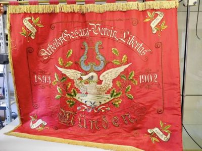 Rote Fahne, auf der "Arbeitergesang-Verein Libertas. Münden. 1893 1902" aufgestickt ist. In der Mitte ein Schwan mit geöffneten Schwingen unter einer Leier, umgeben von Eichenzweigen.