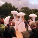 Auf einer Freilichtbühne stehen vier Mädchen in weißen Kleidern mit quallenförmigen Schirmen. Die Bühne ist von Zuschauern dicht umringt.