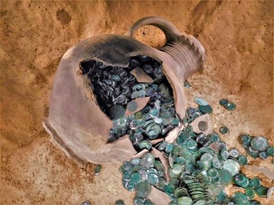 Ein brauner Tonkrug liegt auf der Erde, er ist aufgebrochen und eine große Menge grüne verfärbter, antiker Münzen ist fällt heraus