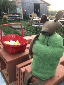 Im Vordergrund steht eine Maus in grünem Kleid als Handpuppe, daneben ein roter Korb. Im Hintergrund sieht man den Garten und die Remise auf dem Meierhof.