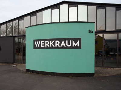 Vor dem Eingang eines verglasten Gebäudes steht eine geschwungene grüne Wand, daran in Großbuchstaben das Wort "Werkraum"