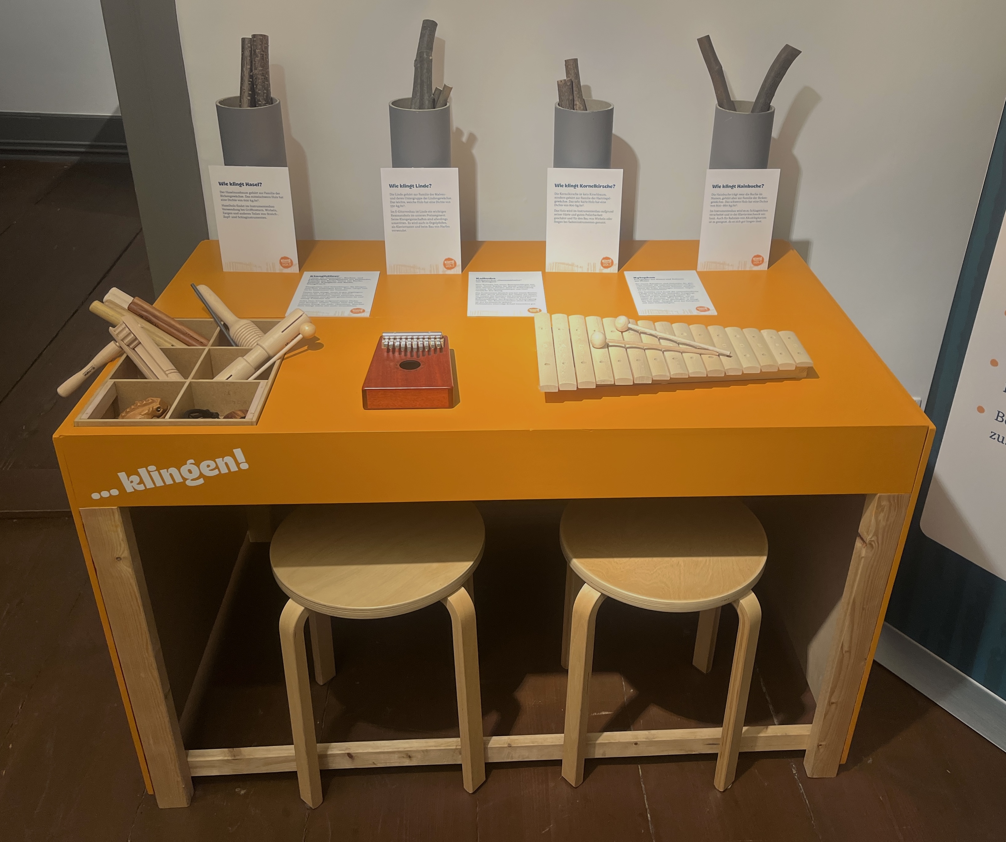 Auf einem orangefarbenen Tisch befinden sich verschiedene Holzinstrumente, wie Klanghölzer, ein Xylophon, eine Kalimba eine Ratsche und ein Klangfrosch. Zwei Hocker stehen vor dem Tisch, rechts daneben steht ein Ausstellungstext.