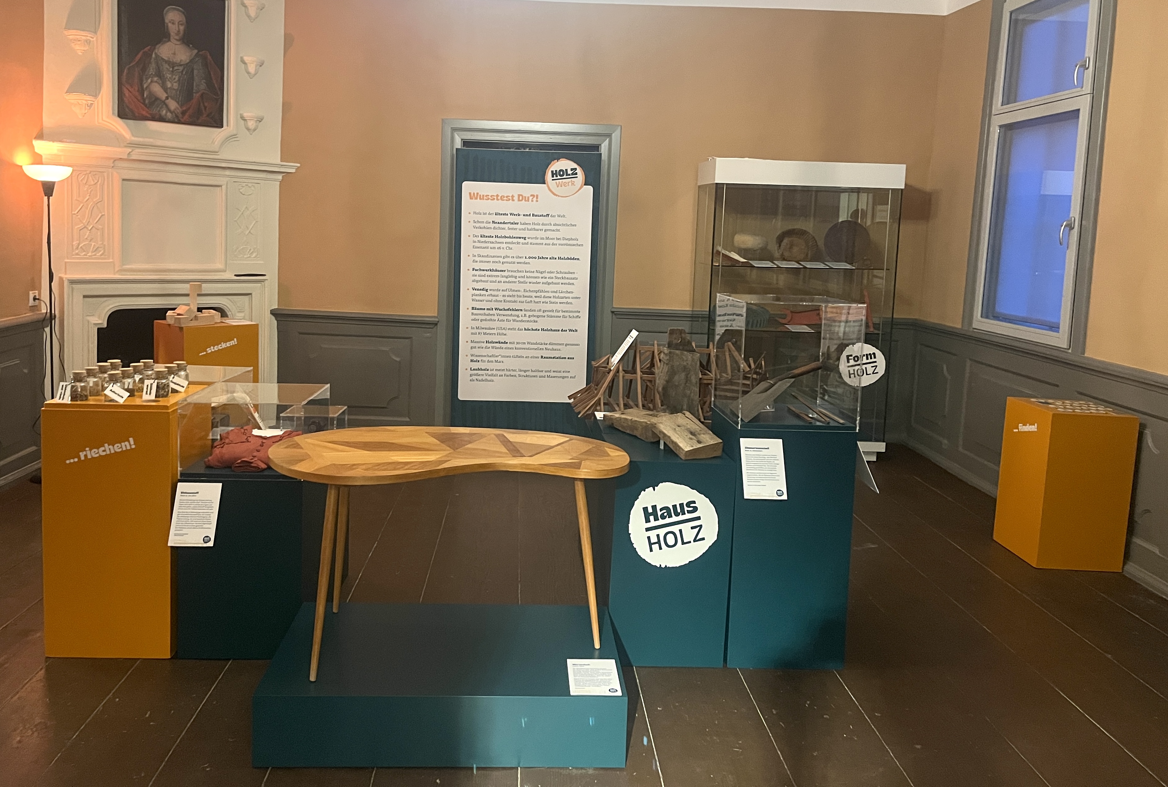 Auf dunkelgrünen und orangenen Podesten stehen verschiedene Holzobjekte, darunter ein Nierentisch, ein Modell eines Dachstuhls, ein Viskosekleid, ein Teufelsknoten. Im Hintergrund steht eine Vitrine mit weiteren und Objekten und ein Ausstellungstext.