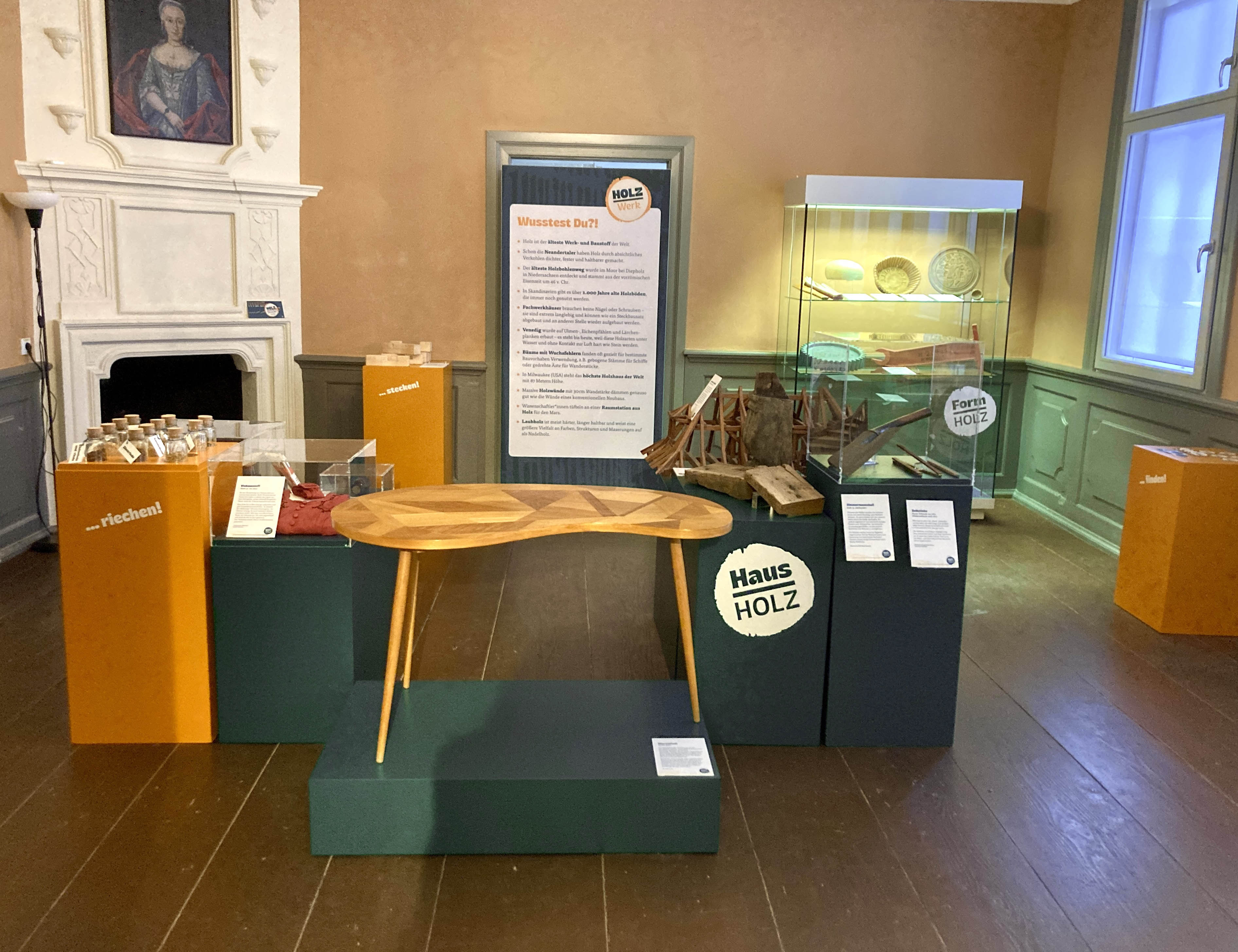 Blick in einen historisch wirkenden Raum mit orangenen und dunklen, grünblauen Podesten mit Exponaten darauf. Im Vordergrund ein Nierentisch, dahinter u.a. ein Dachmodell. Rechts im Hintergrund weitere Objekte in einer Vitrine und ein Ausstellungstext.