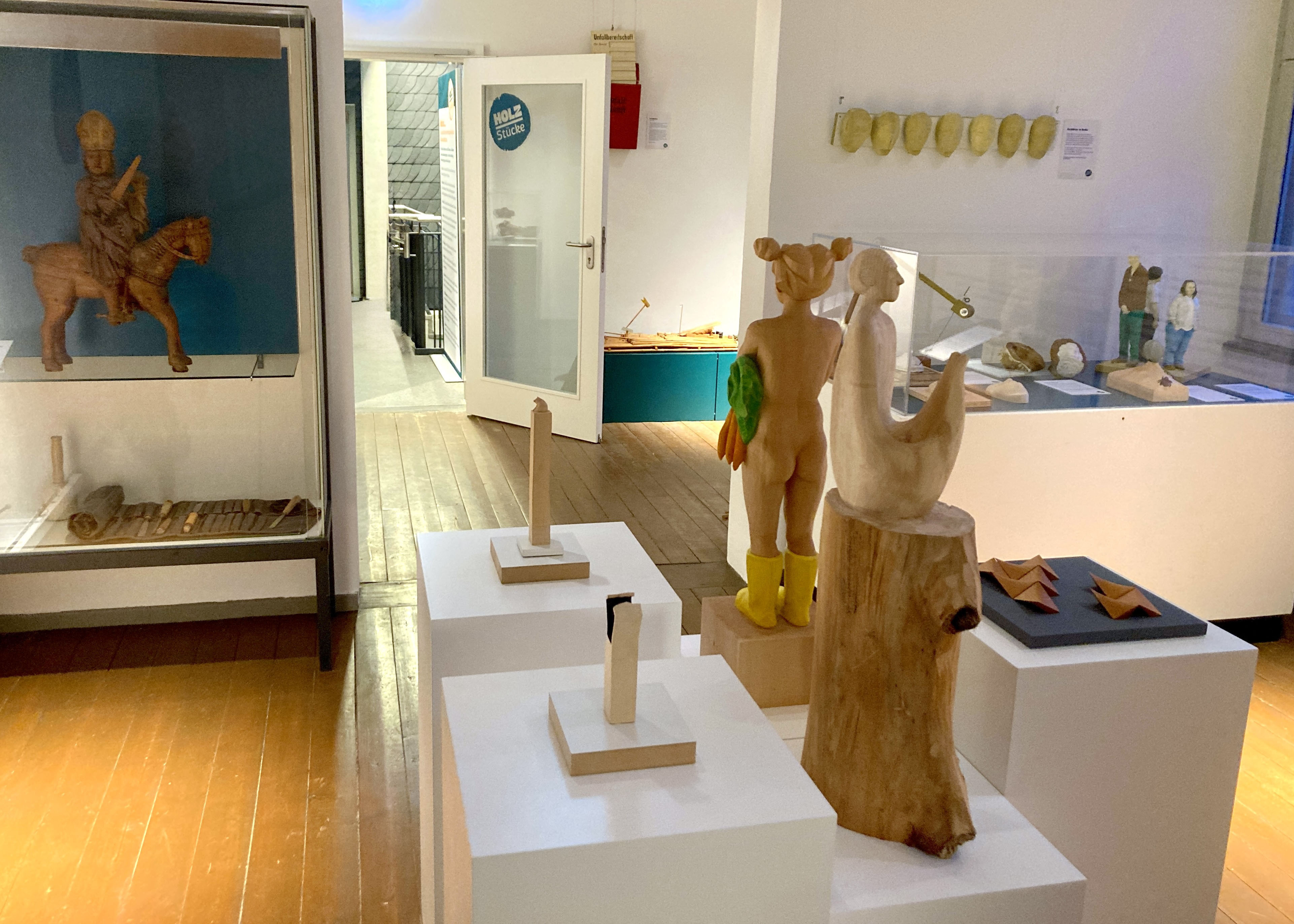 Blick in einen Ausstellungsraum mit weißen Podesten, auf denen Holzskulpturen stehen. Dahinter zwei Vitrinen mit weiteren Holzkunstwerken und Werkzeugen. Im Hintergrund eine geöffnete Tür mit Aufschrift: HolzStücke.