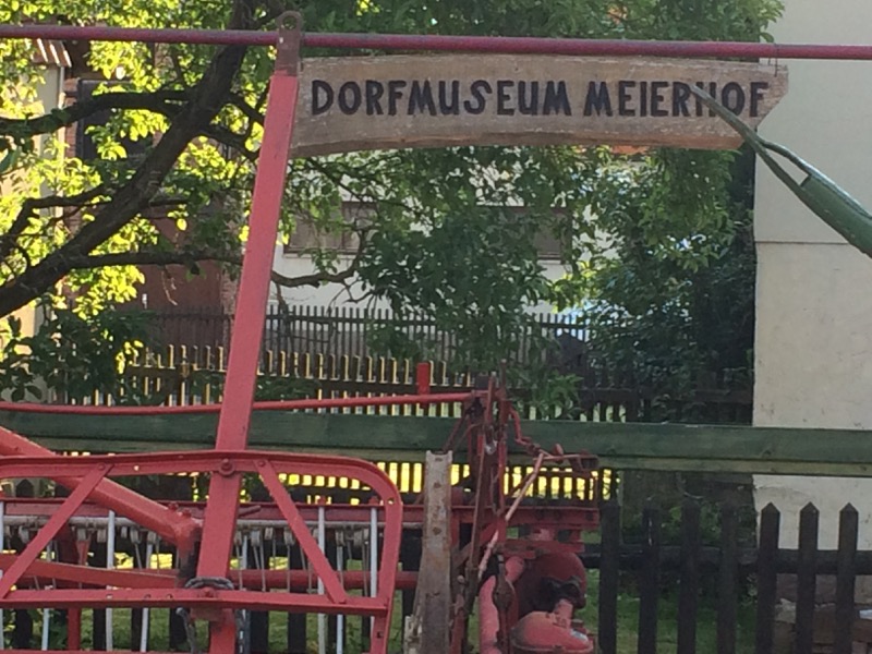 An einem rot gestrichenen landwirtschaftlichen Gerät hängt ein Holzschild mit der Aufschrift "Dorfmuseum Meierhof"