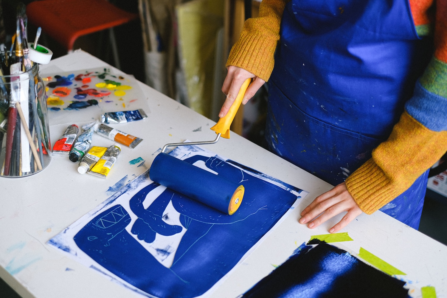 Eine Person mit blauer Schürze über einem bunten Pullover rollt eine Farbwalze mit blauer Farbe über einen Linolschnitt. Auf der Arbeitsplatte liegen Farbtuben, daneben ein Glas mit Pinseln.