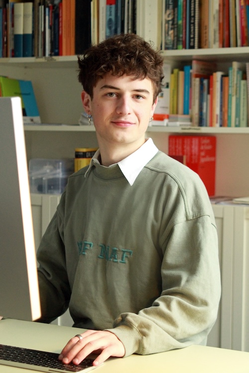 Portrait eines jungen Mannes, der an einem Schreibtisch sitzt und freundlich in die Kamera lächelt. Er hat helle Haut, kurze dunkle Locken und trägt ein blassgrünes Sweatshirt. Hinter ihm sieht man einen Teil eines Bücherregals.