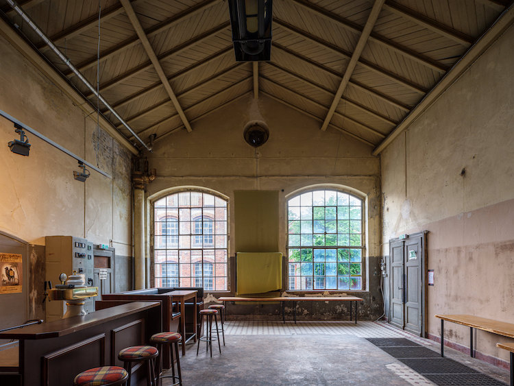Aus einer alten Fabrikhalle schaut man durch zwei große Fenster nach draußen. Links erkennt man eine Bar.